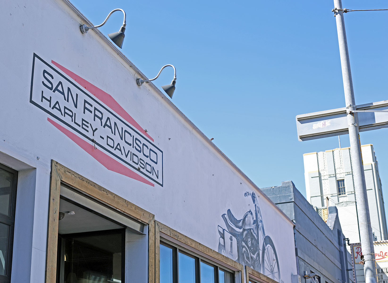 A white building with a "San Francisco Harley-Davidson" sign and a mural of a Harley Davidson motorcycle on the side wall under a clear blue sky.