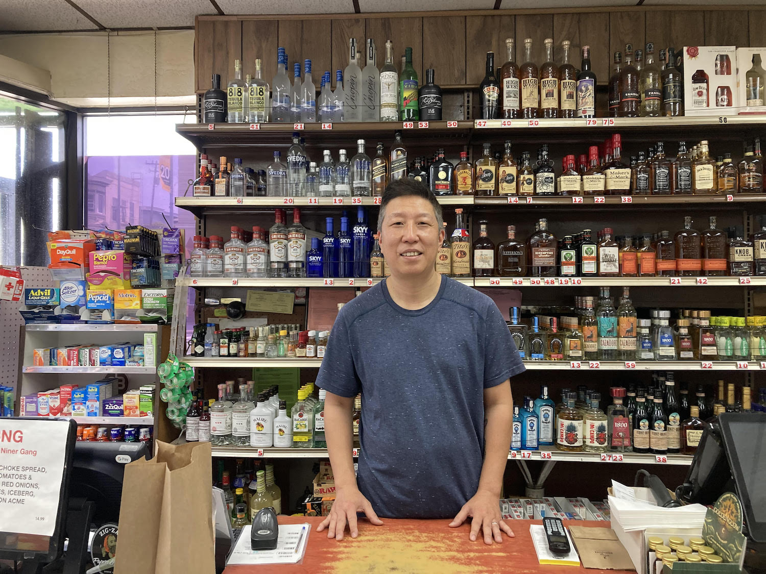 A man stands behind the counter of a mart, with shelves filled with various bottles of alcohol and other products in the background.