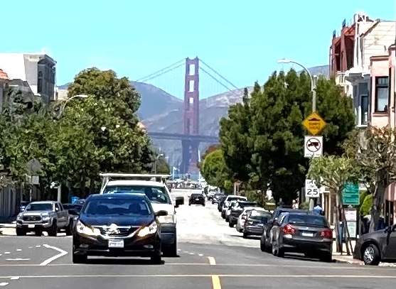 A street view showing the Golden Gate Bridge in the distance, with cars parked along the sides and trees lining the road on a clear day.