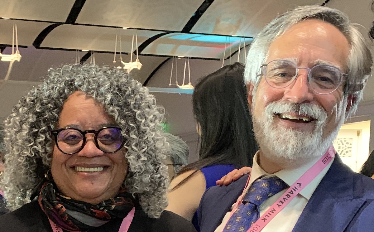 Two adults are smiling at an indoor event. The woman on the left has curly gray hair and glasses. The man on the right has white hair, a beard, and glasses, and is wearing a suit and tie.
