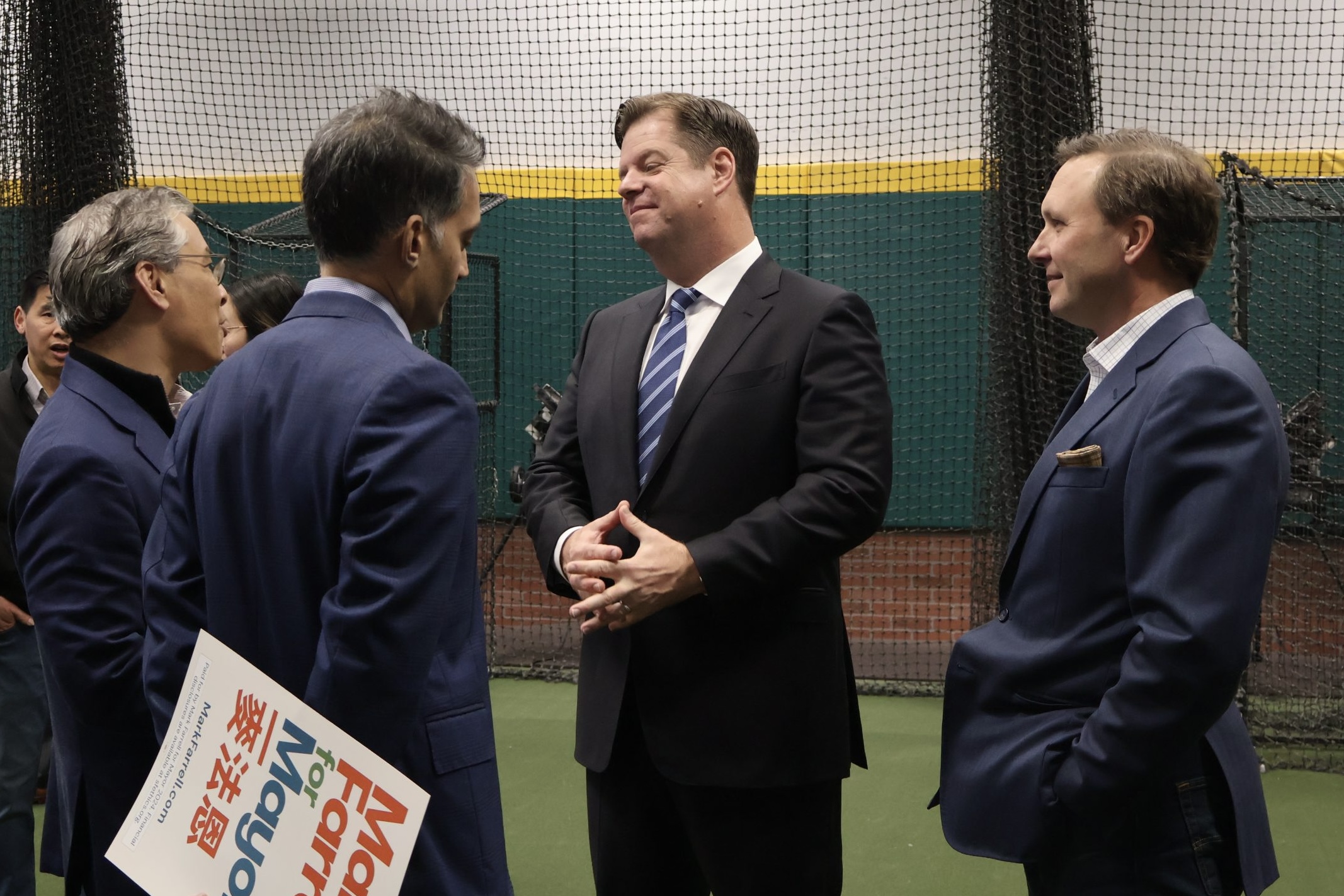 Four men in business attire are conversing in an indoor setting, with one holding a sign. A netted wall and green flooring are visible in the background.