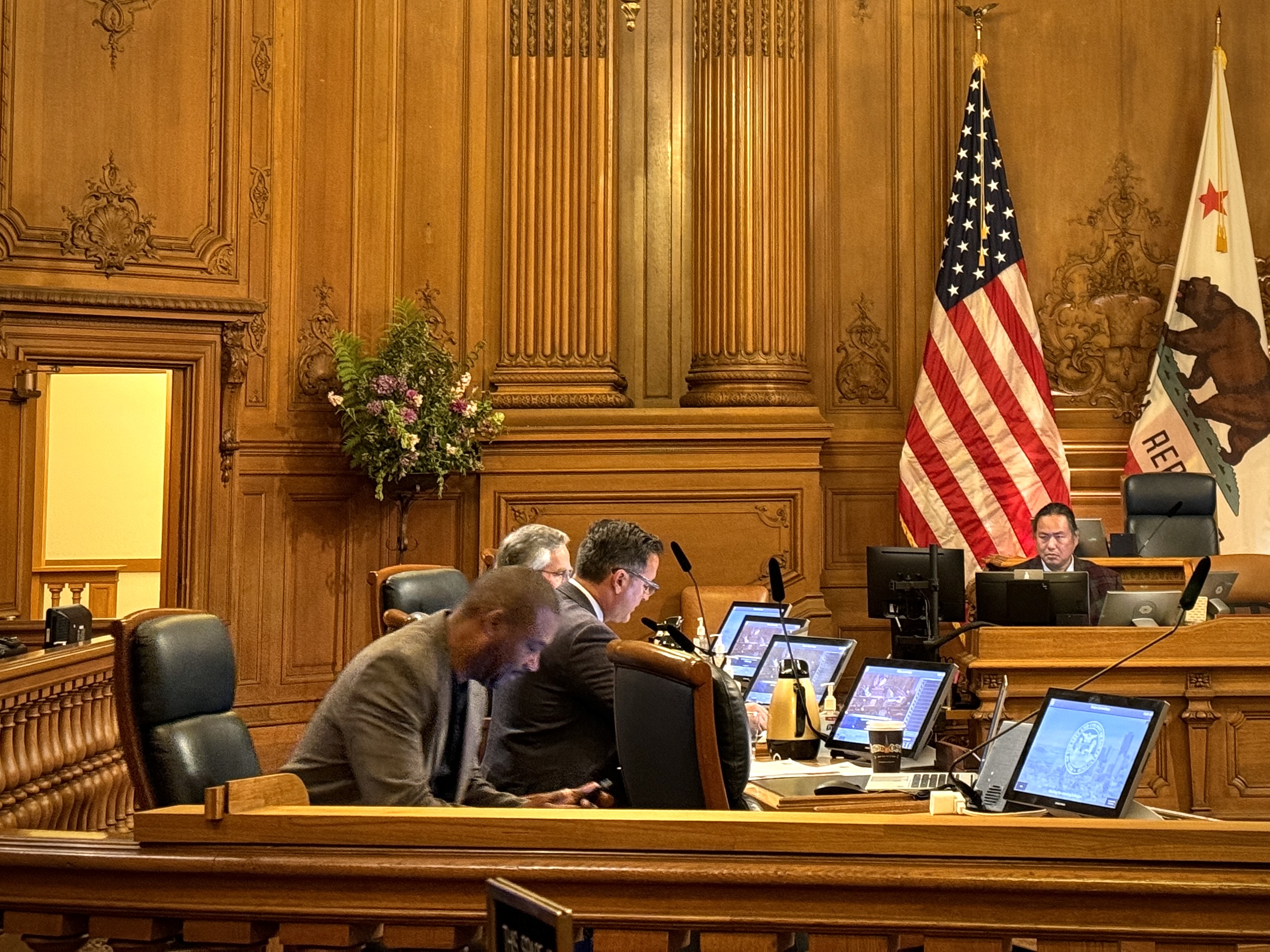 Four individuals sit at a wooden desk with multiple computer monitors in a room with ornate wood paneling, an American flag, and a California state flag.