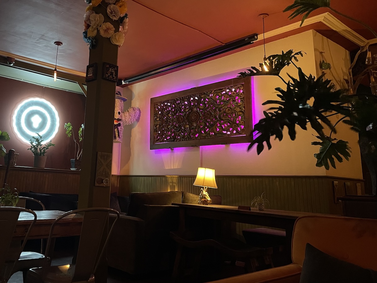 A dimly lit room with eclectic decor featuring a neon light, ornate wall art with purple backlighting, various plants, a lamp on a table, and comfortable seating arrangements.