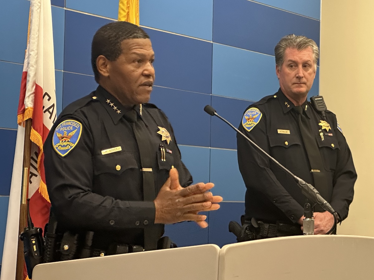 Two police officers in uniform are standing at a podium; one is speaking while the other stands by.