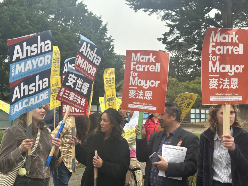 A group of people holding campaign signs for Ahsha Safaí, Mark Farrell, and Breed, all running for mayor. Some signs feature both English and Chinese text. Trees and an overcast sky are visible in the background.