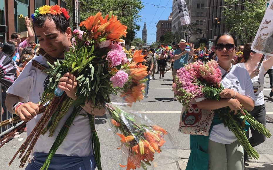 Two individuals carrying large bouquets of flowers and wearing flower crowns walk in a street parade. The parade is set in an urban environment with people and buildings in the background.