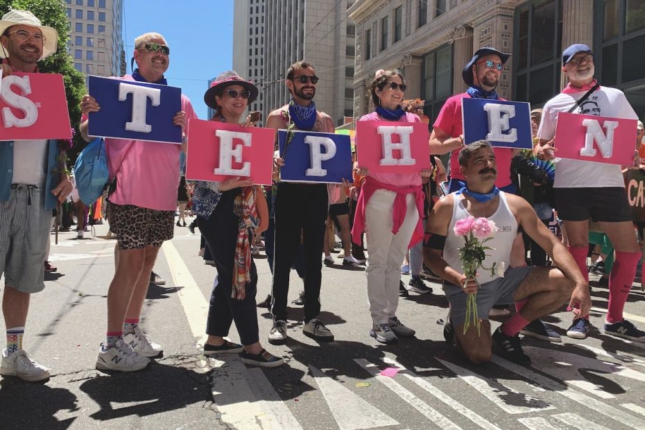A group of people stands on a city street holding large letters that spell "STEPHEN." One person kneels in front, holding a bouquet of pink flowers.
