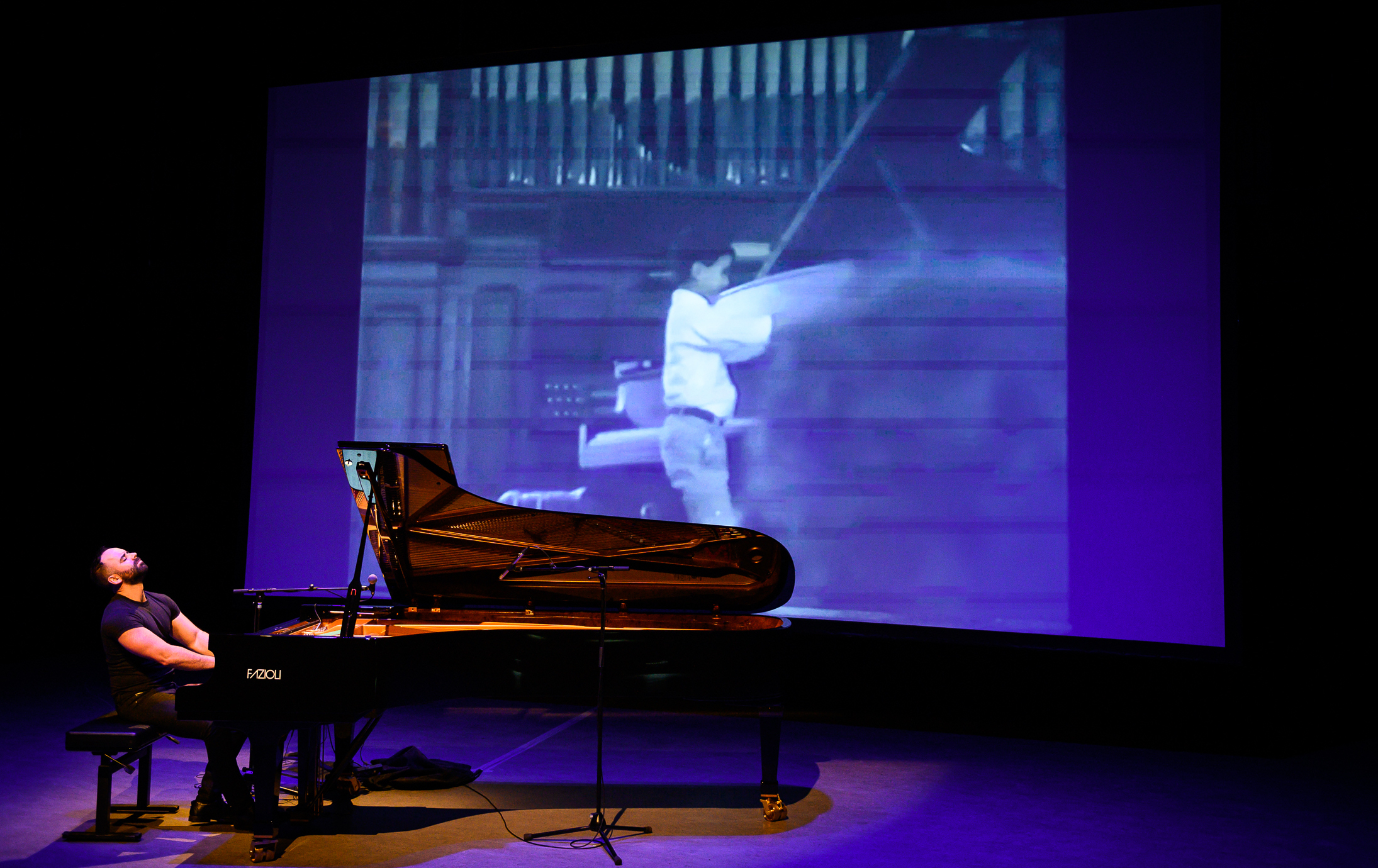 A musician plays a grand piano on stage, while behind him, a large black-and-white image of a person swinging an axe is projected on a screen—a true showcase of Adam Tendler's flair for experimental music.