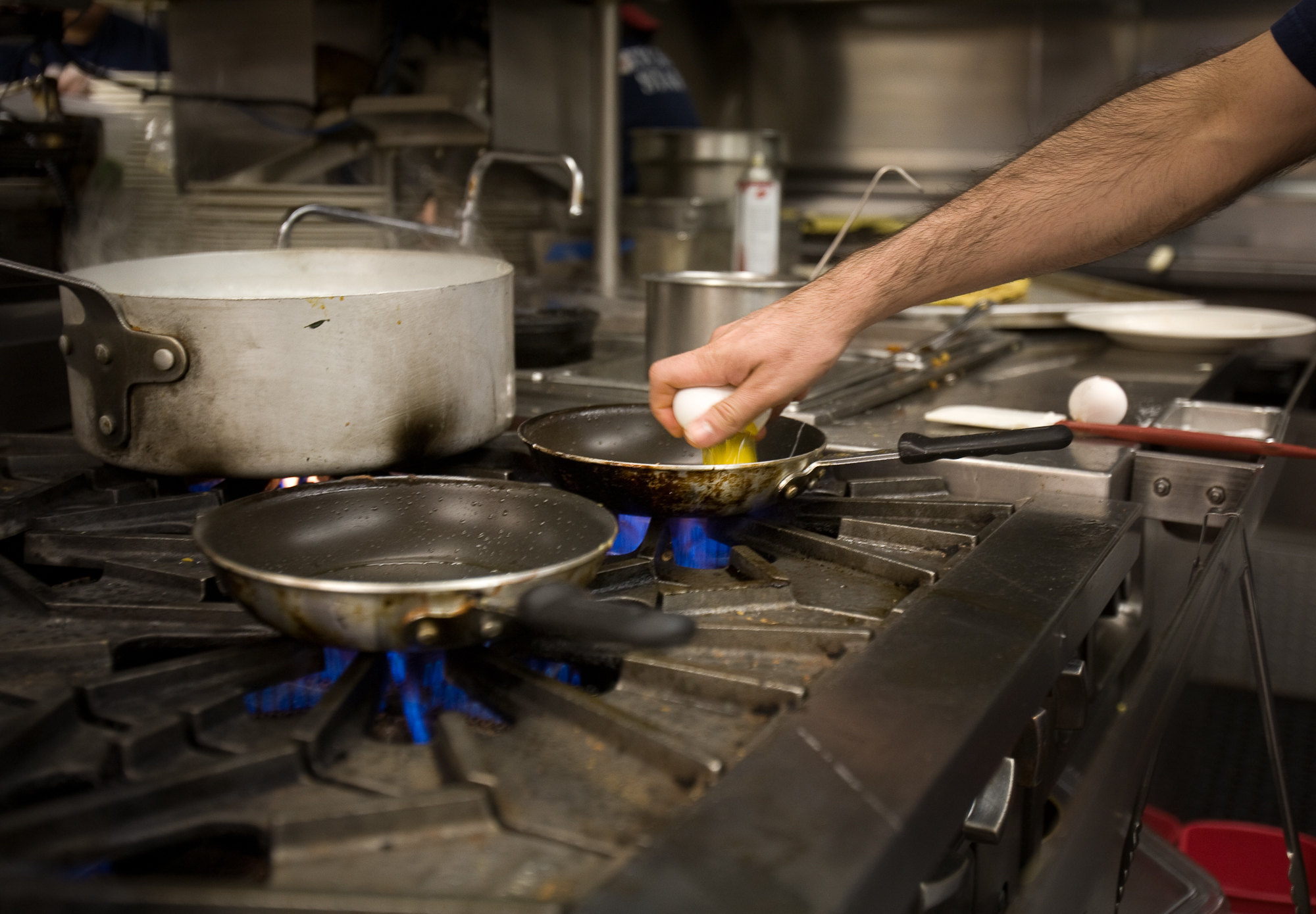 A chef cracks an egg over a pan on a gas stove.
