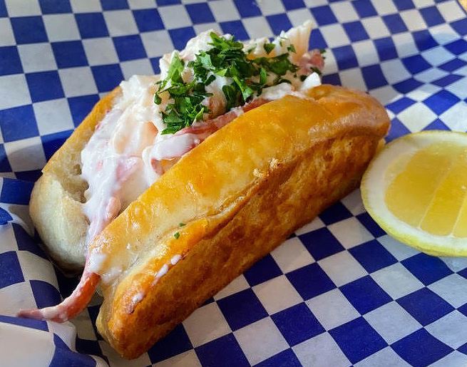 A lobster roll with a lemon wedge next to it sitting on blue and white checkered paper
