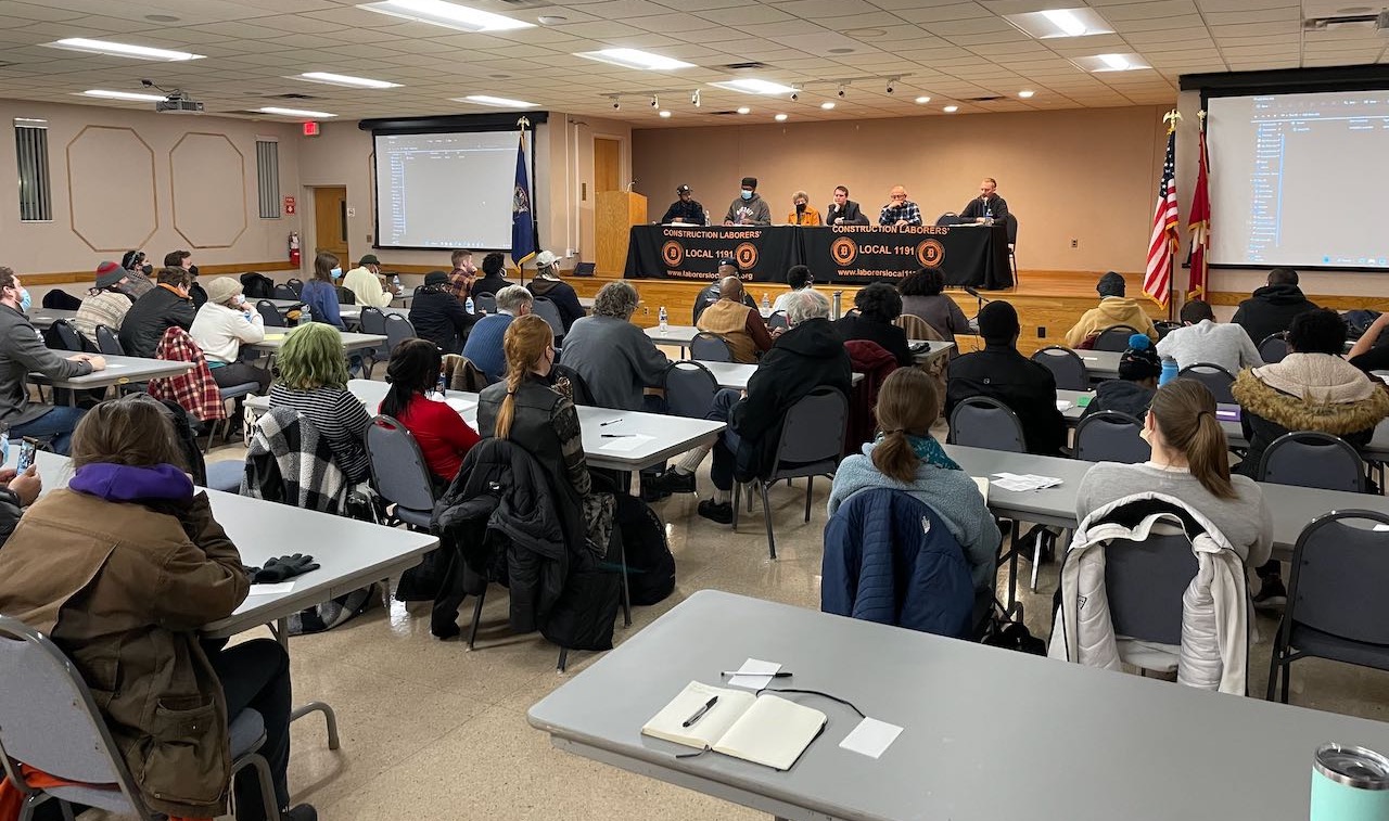 Large audience seated in a meeting hall, in front of six people on a stage seated at a table with microphones. The table is draped in black cloth with yellow text that says, “Construction Laborers’ Local 1191. www.laborersinternational.com.“