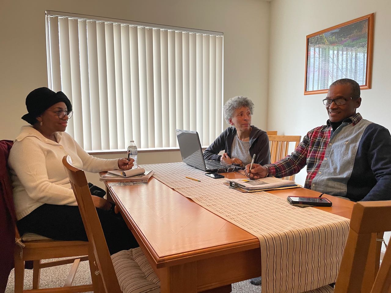 Three people seated around a wooden dining table writing notes and talking. From the left to right: a dark-skinned person with a black hat, glasses and pink sweater; a dark-skinned person with grey hair and a grey hoodie; and a dark-skinned person with black hair and a patterned shirt.