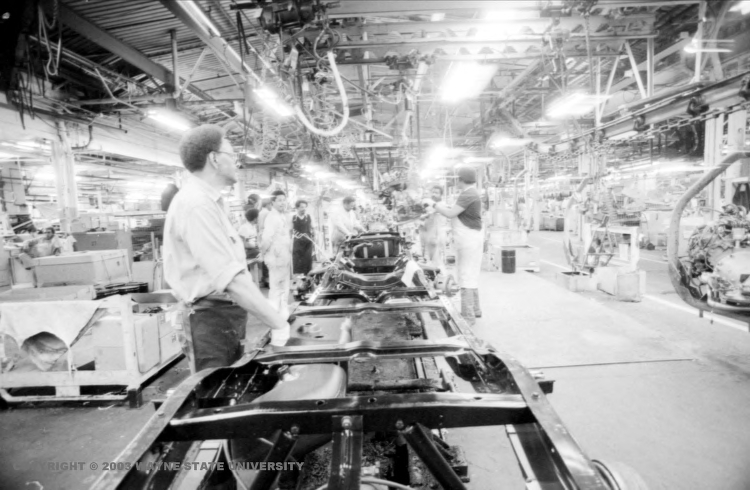 Bright lights shine in a black-and-white photo of an automotive factory as multiple men work on the assembly line.