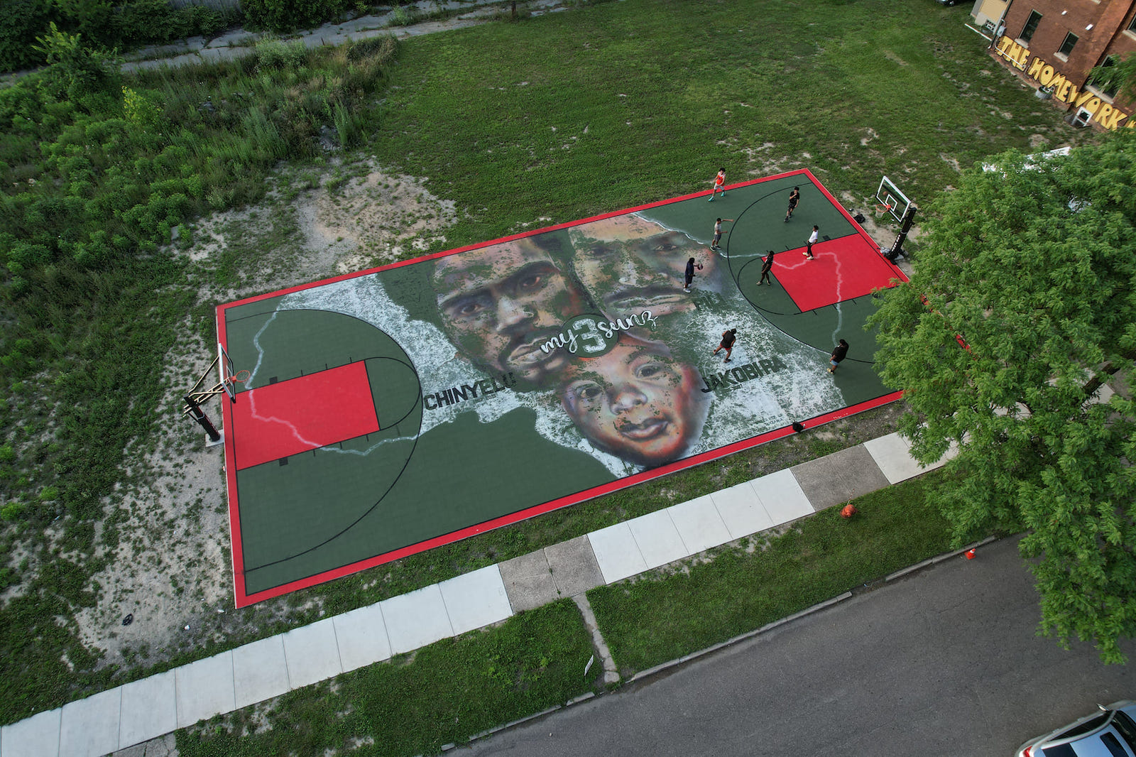 Green-and-red basketball court in a grassy field with people near the right basketball hoop. In the center of the court is a drawing of two Black men, a Black child, and text that says, “My 3 Sunz. Chinyeli. Jakobi RA. Pili.”