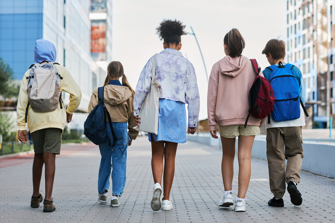 Five students walk along a street wearing backpacks on an overcast day.