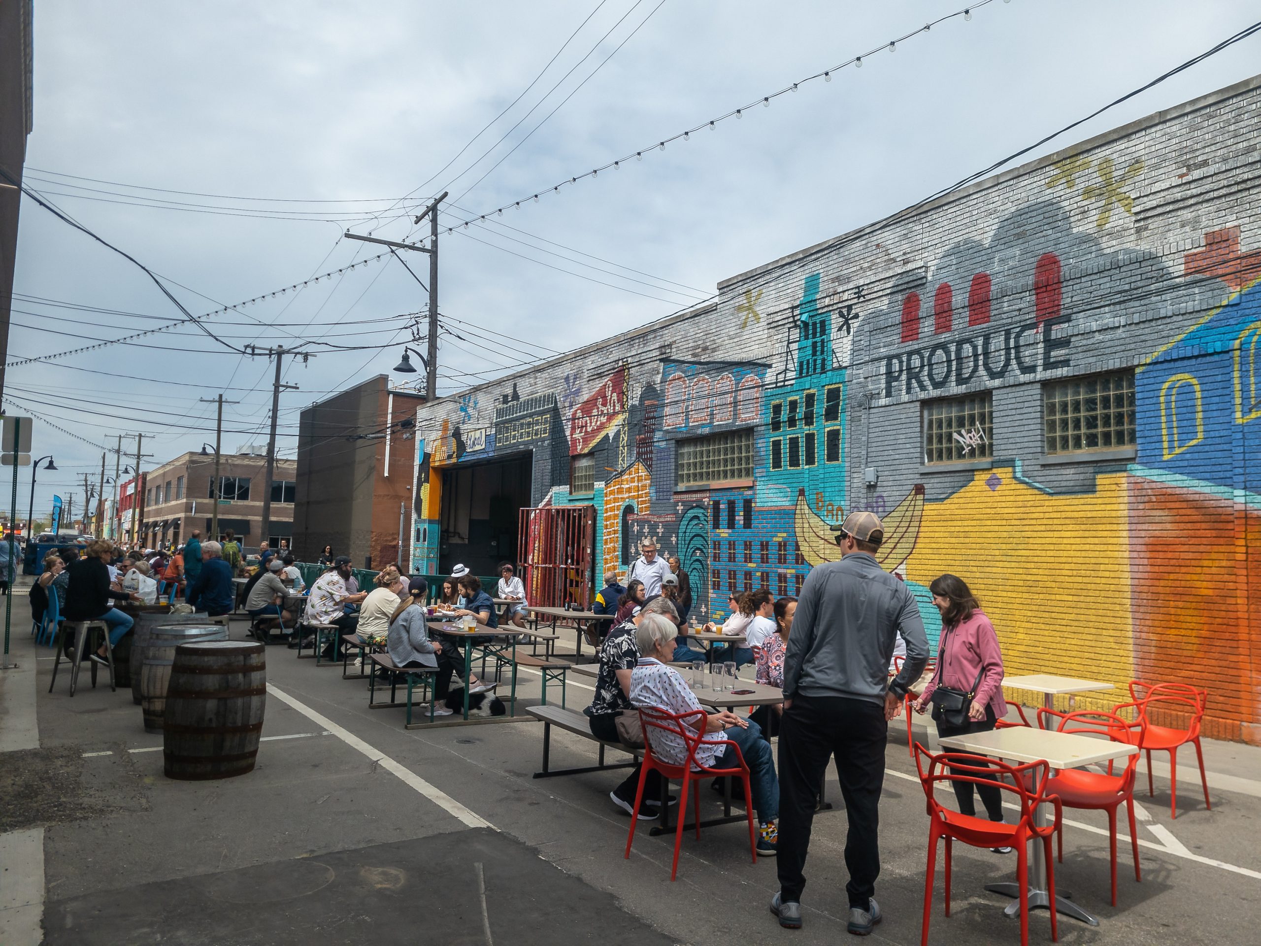 Many diners seated outdoors at tables set up in a narrow street, next to an old brick industrial building with a colorful mural of a cityscape with the words “fresh” and “produce.”