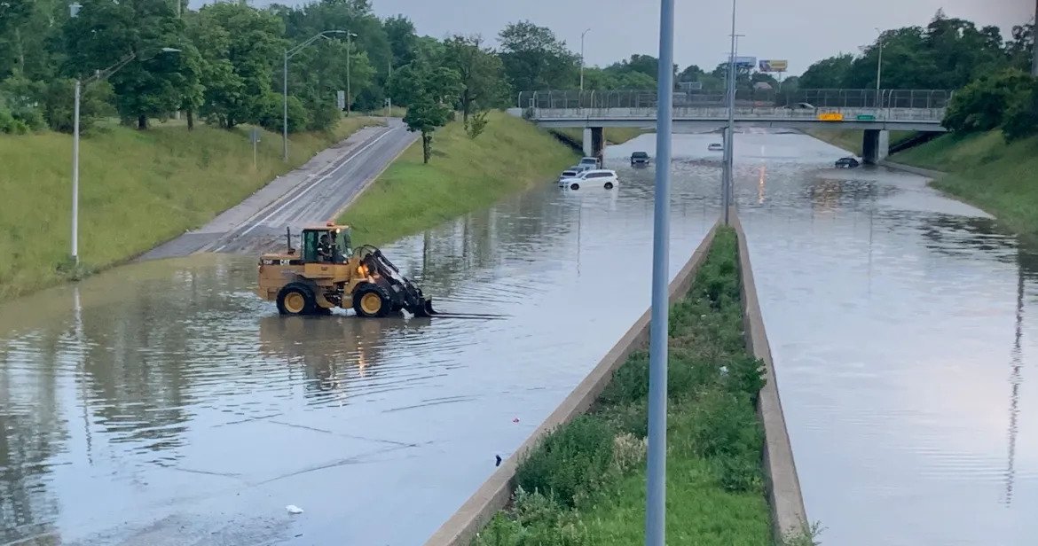 Water fully covering highway lanes and rising slightly onto entrance ramp, with a person driving a construction vehicle across lanes. There are several partially submerged cars.