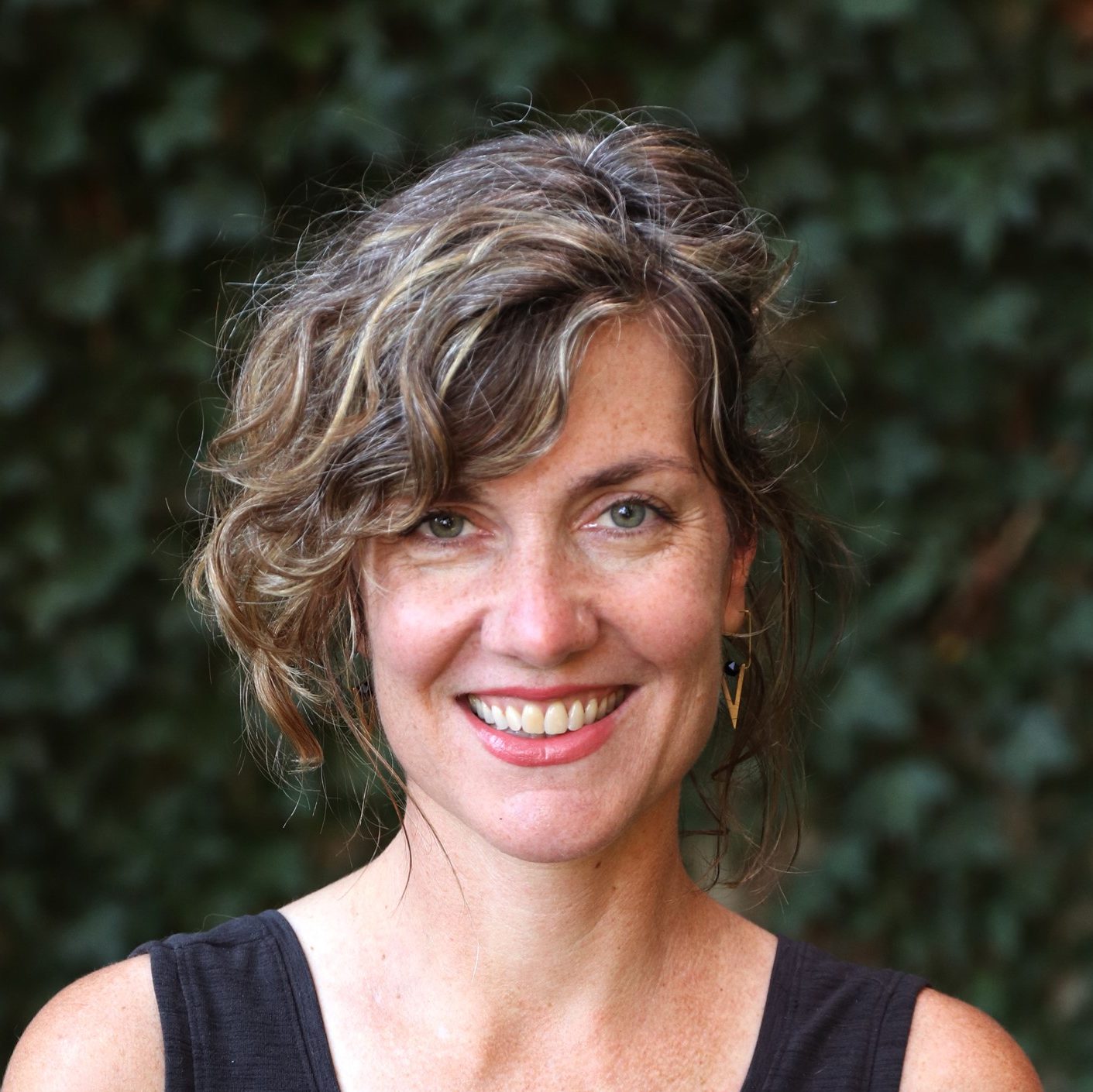 A white woman with chin length wavy hair smiles for the camera in front of a wall covered in ivy. She is wearing a black v-neck tank top