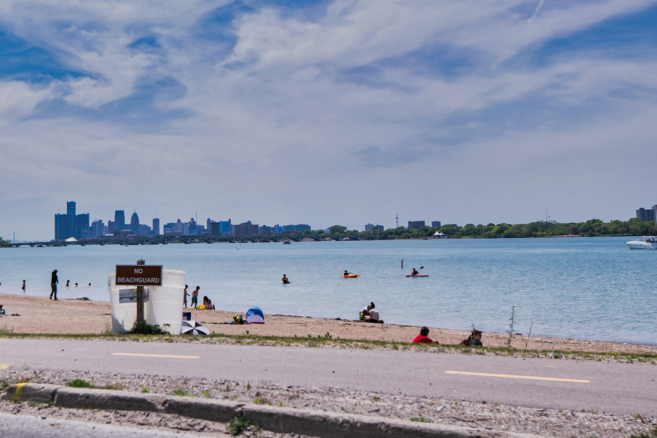 Beach with people swimming, kayaking, walking or sitting in the sand, behind a “No Beachguard” sign and across the water from a city skyline.