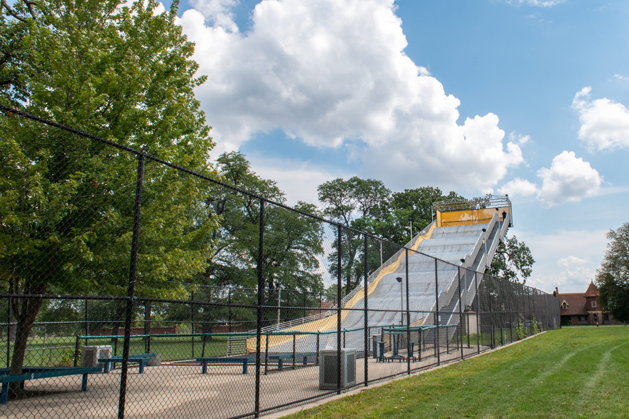 A tall, wide, wavy metal slide with a staircase leading to the top, behind a chain-link fence.