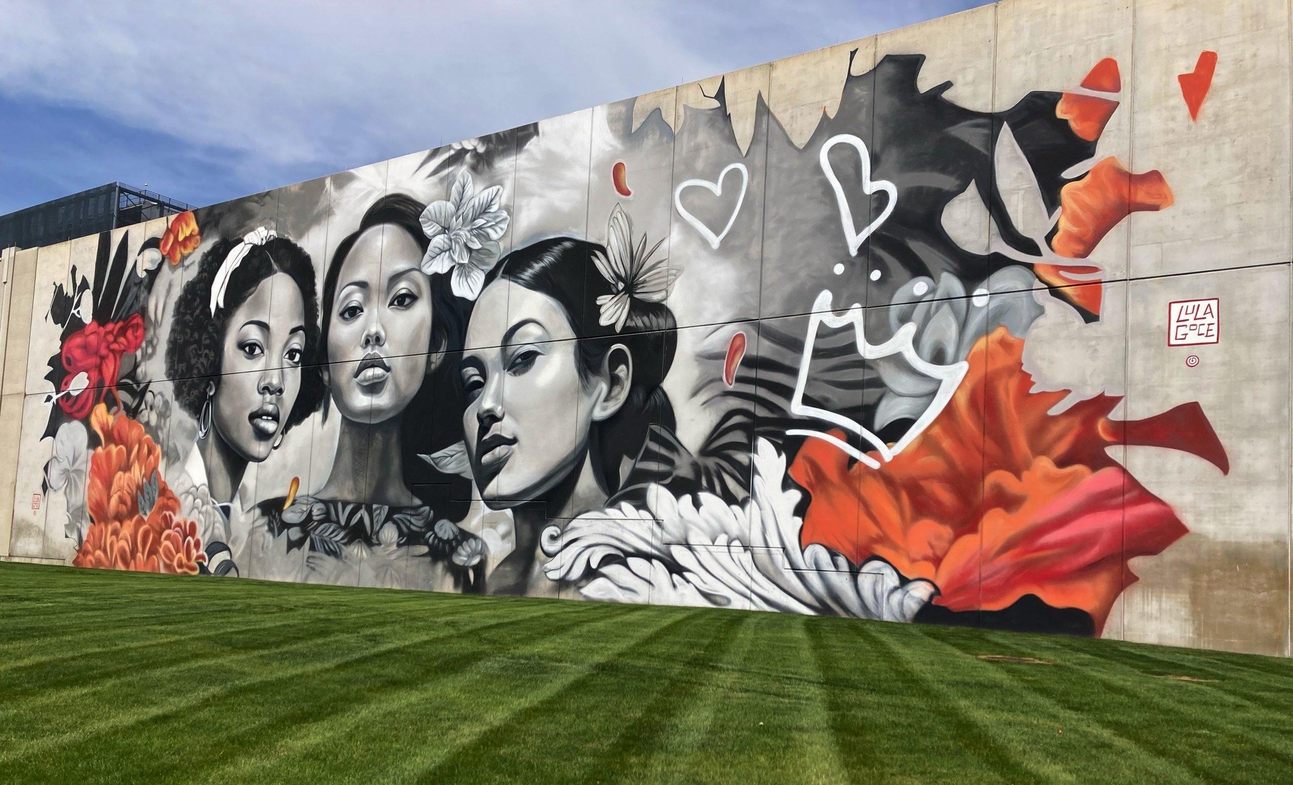 Large-scale mural on the side of a building featuring three women’s faces in black and white with orange flowers to the left and right.