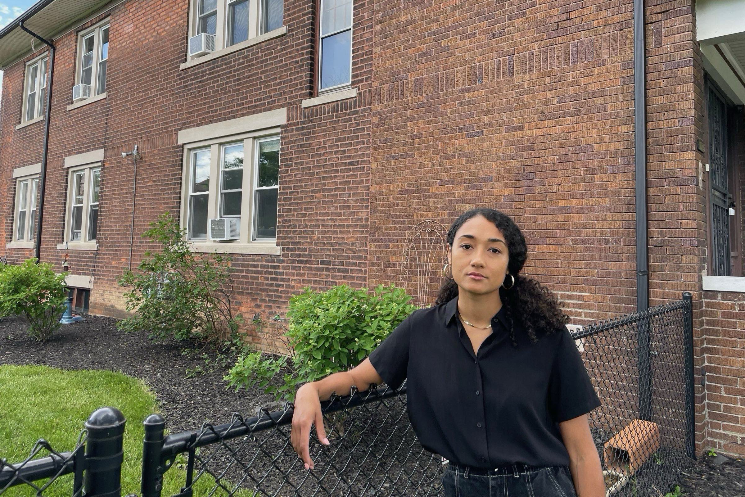 Black woman in black shirt stands outside a brick, two-story house.