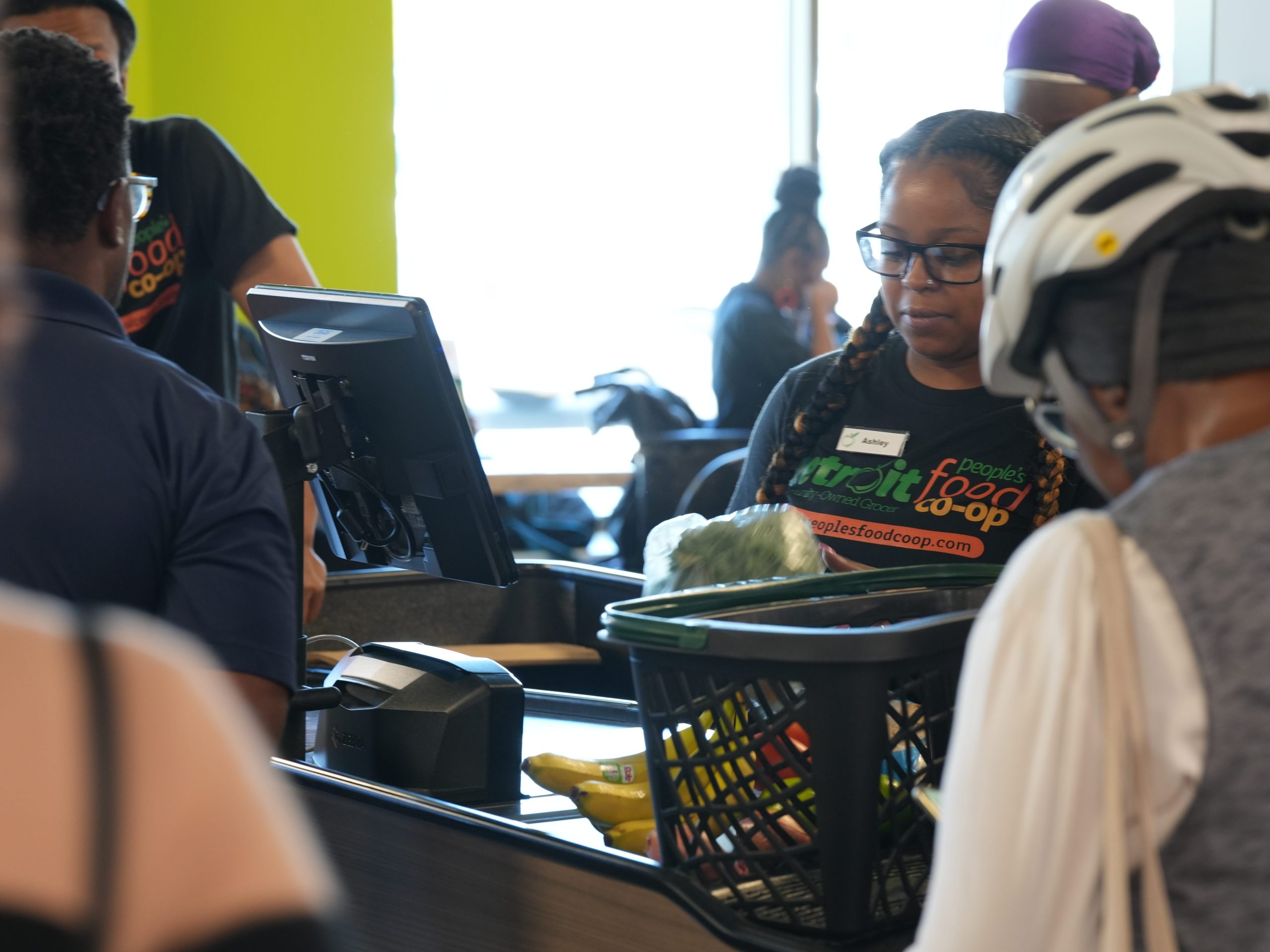 Woman wearing a black shirt that reads “Detroit People’s Food Co-op” rings up groceries at a cash register for a person wearing a white bike helmet