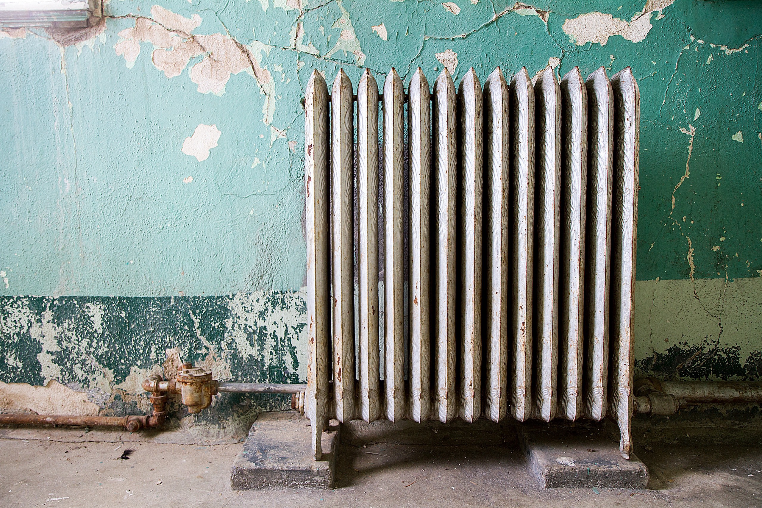 Rusty radiator in front of a cracked wall with peeling aqua-green paint.