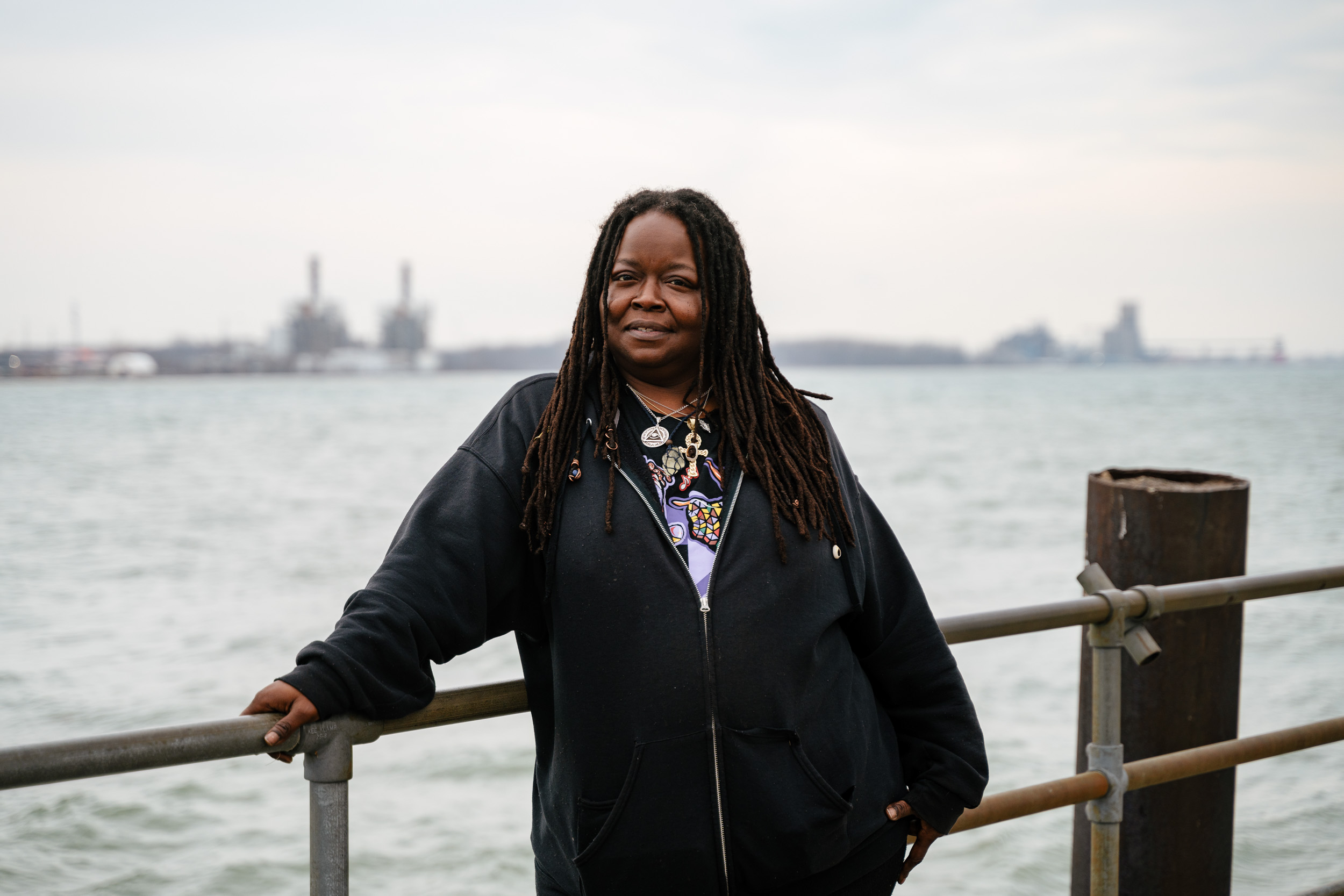 Black woman with locs wearing a black jacket and two large silver pendants stands in front of a river with a city in the distance.