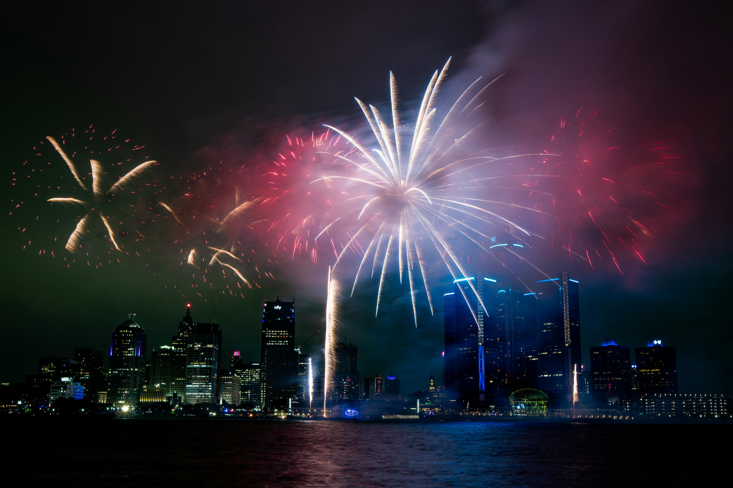 Red and white fireworks going off at dusk over a river in front of a city skyline.