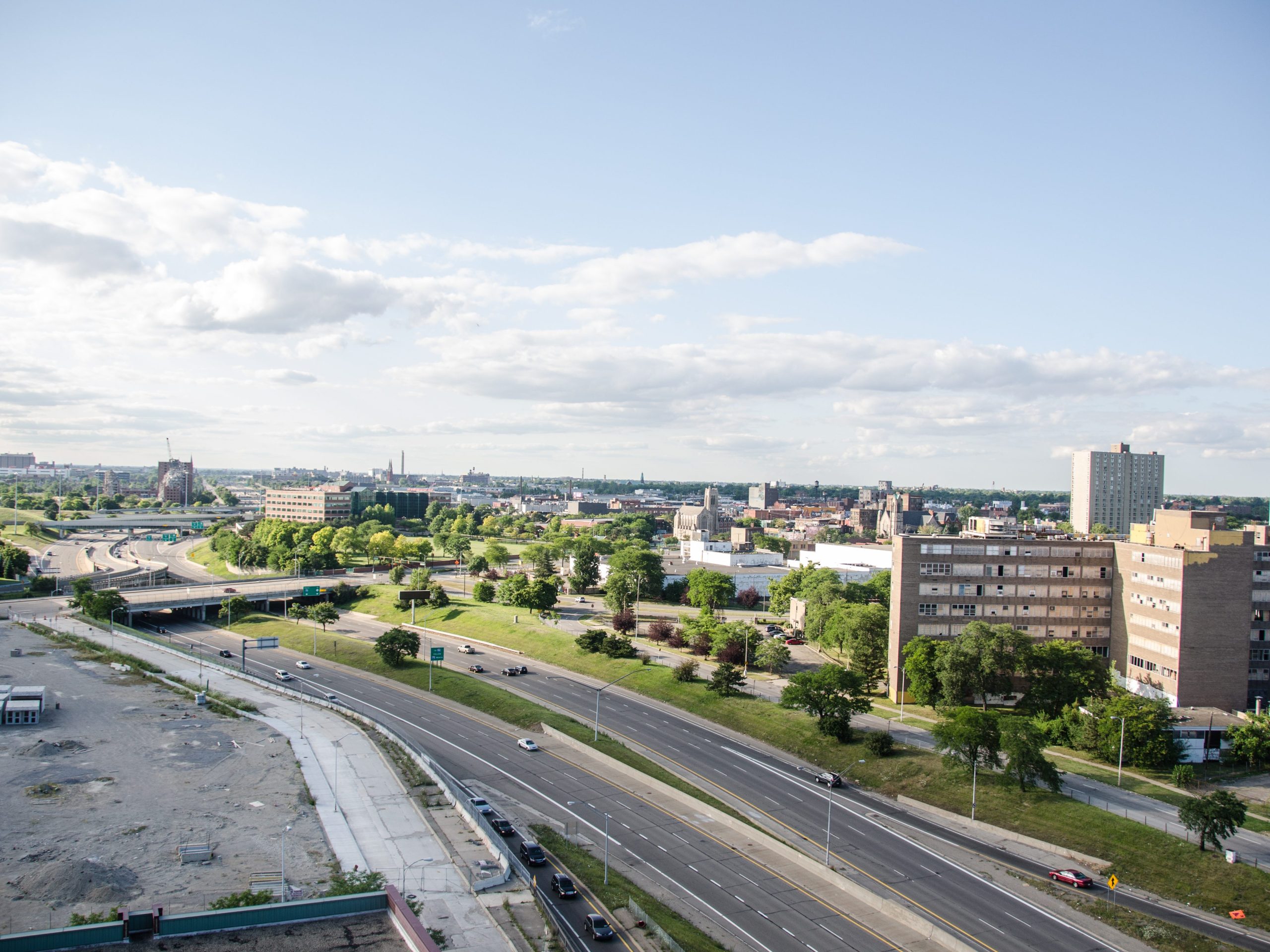 Aerial view of a six-lane freeway in a dense urban area. The roof of a building is on one side; high-rises and trees are on the other side.