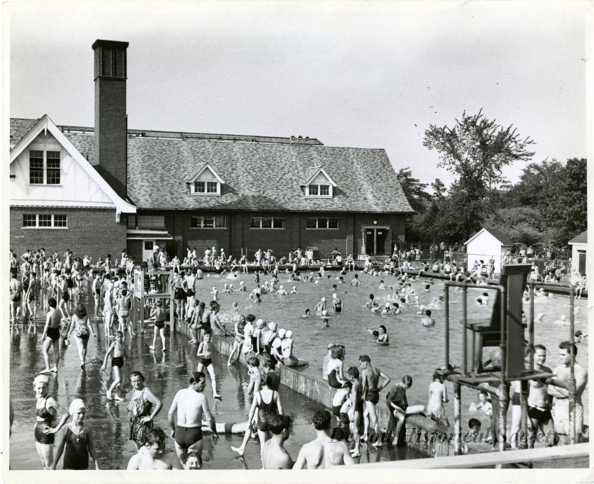 Black-and-white photo of a crowded outdoor pool surrounded by many people in front of a brick building with a gable roof and chimney.