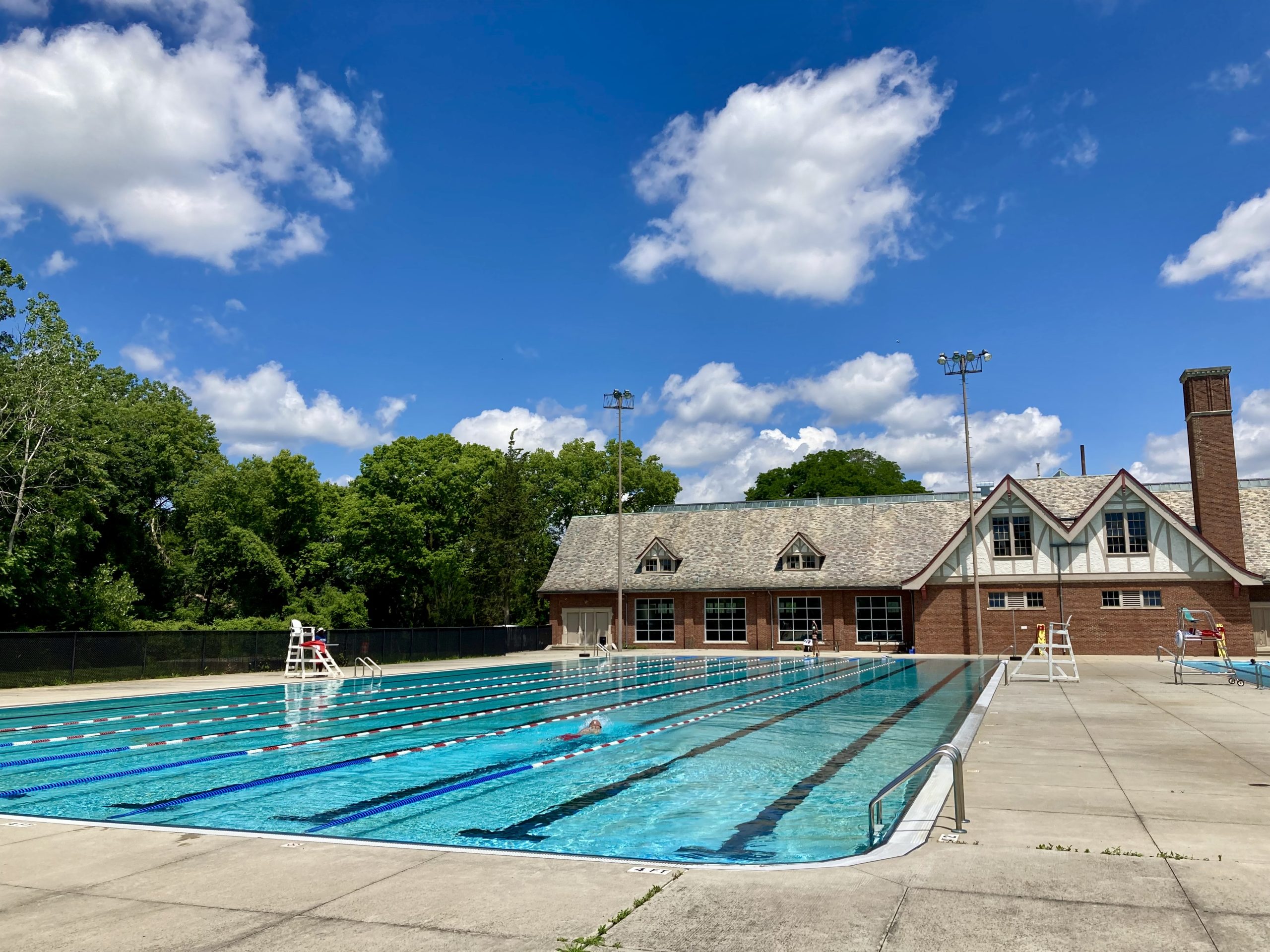 A person swimming laps in an outdoor Olympic-size pool with bright blue water in front of a brick, one-story building with a gable roof and large picture windows.