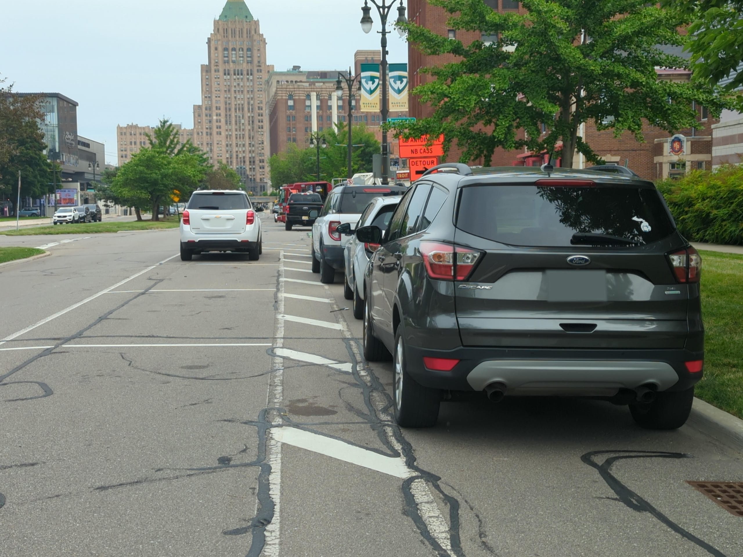 Three cars are parked on a boulevard inside a painted bike lane while a white car sits in a parking space. Down the street, a black pickup truck and a red fire truck also block the bike lane. In the distance is an Art Deco skyscraper with a green roof.