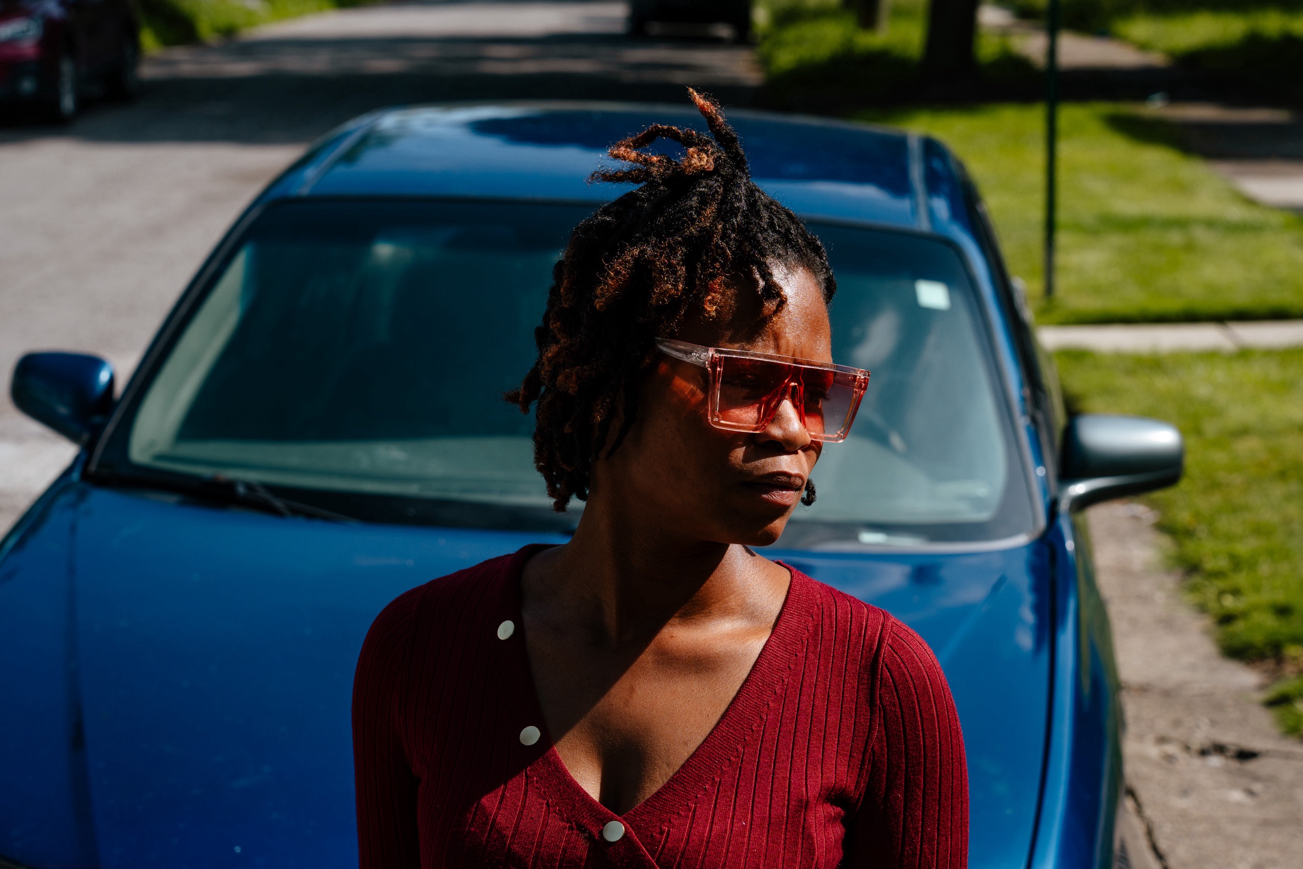 A Black woman wearing a red dress and sunglasses leans against the hood of a blue car parked in the street while the sun shines on her.