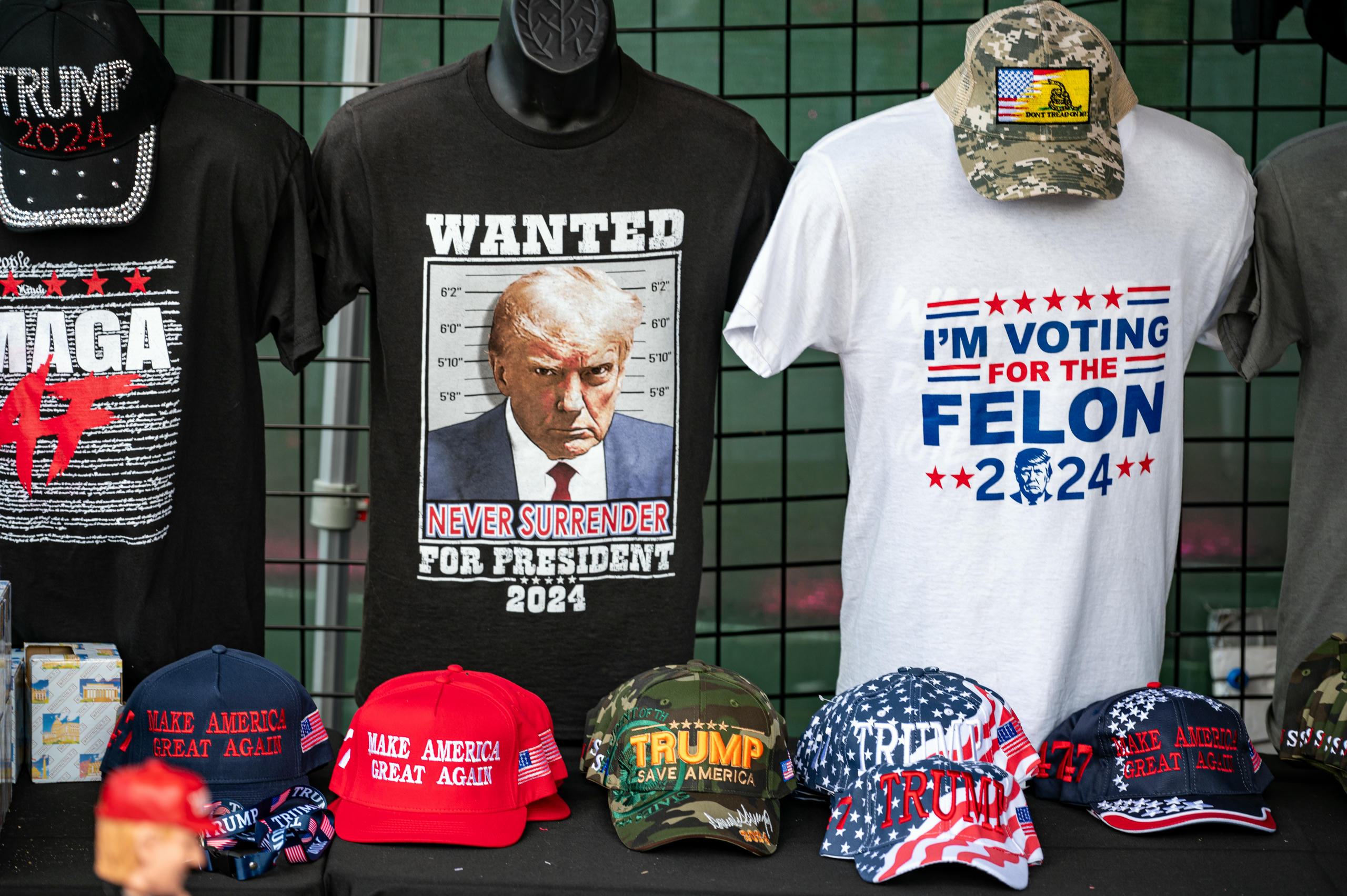 A merchandise table with three T-shirts for sale. One features a mugshot of former President Trump on a black background. The t-shirt to the right has a white background and “I'm voting for the felon, 2024” printed in blue and white ink.