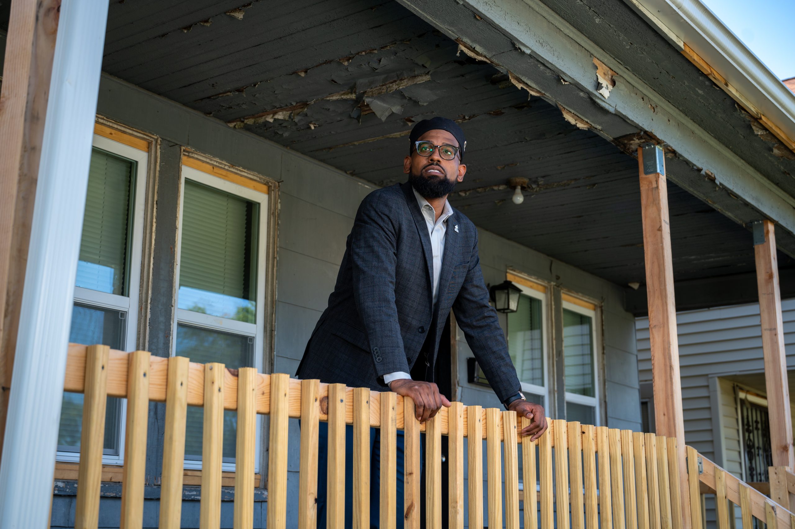 A man wearing a suit and glasses stands on the porch of an older house with peeling paint.