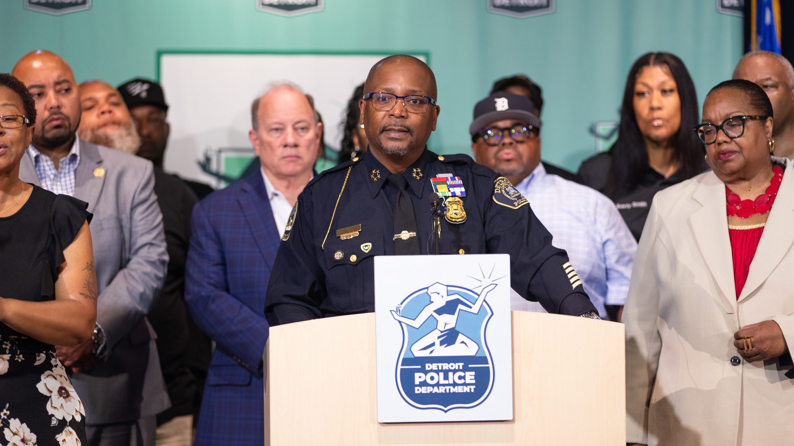 Black man wearing a police uniform stands at a podium with a crowd of people behind him in front of a green backdrop with the City of Detroit logo.