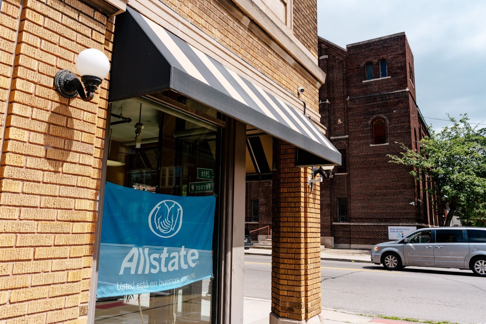 A blue flag that says, in Spanish, “Allstate, you’re in good hands.” The flag is in the window of a yellow-brick storefront next to an urban street with a passing car. In the reflection are street signs that say “Vernor W” and “25th.”