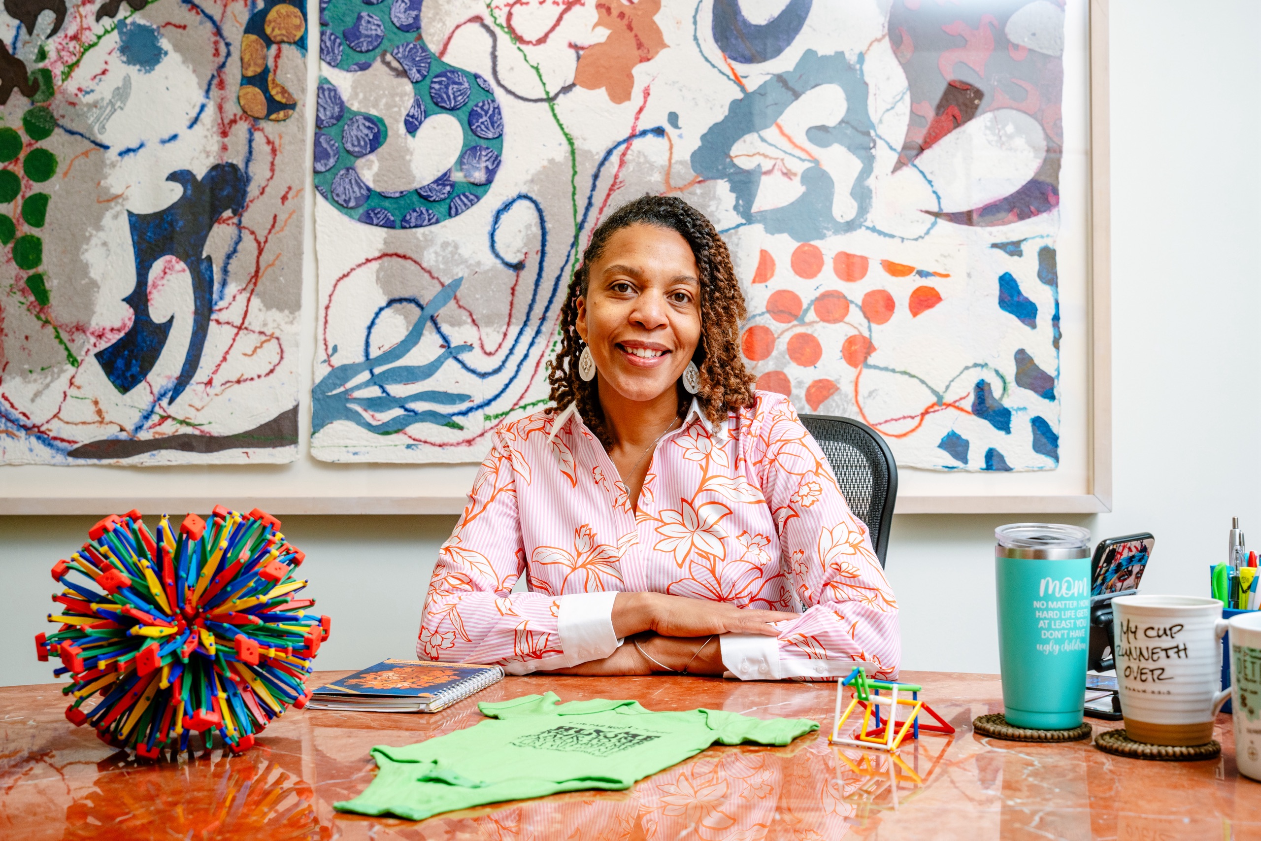 A Black woman wearing a pink shirt smiles in a bright office with a colorful artwork behind her