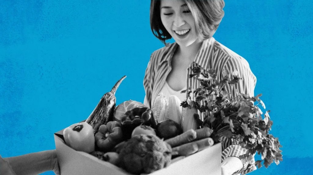 Adult female, smiling, holding a box of fresh fruit and vegetables after receiving a delivery from a produce delivery service