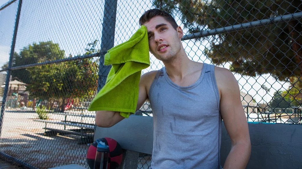a person is standing by a fence wearing a gray tank top and wiping sweat with a towel
