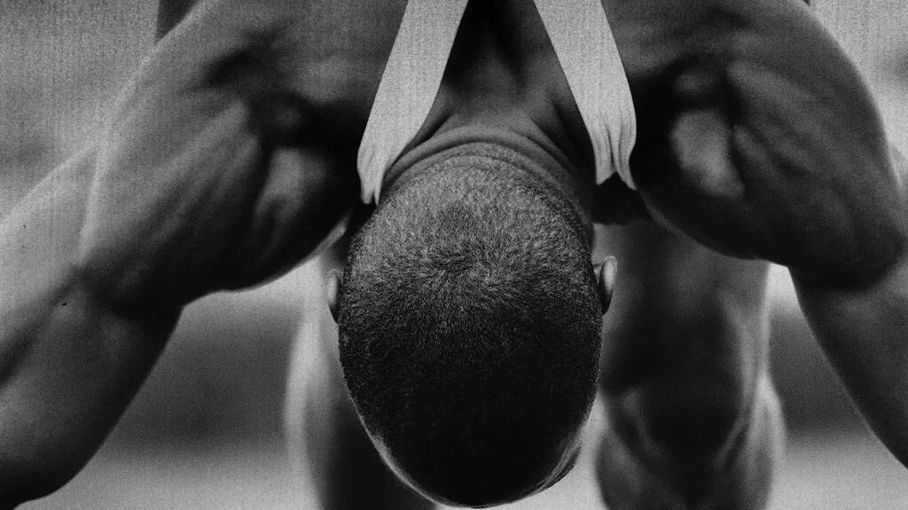 Black-and-white photo of an athlete bent over as they prepare for a sprint. Their muscles look rigid.