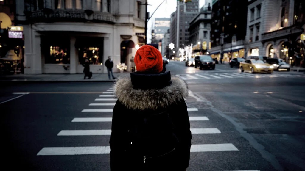 A person about to cross a city street wearing a winter coat and red hat.