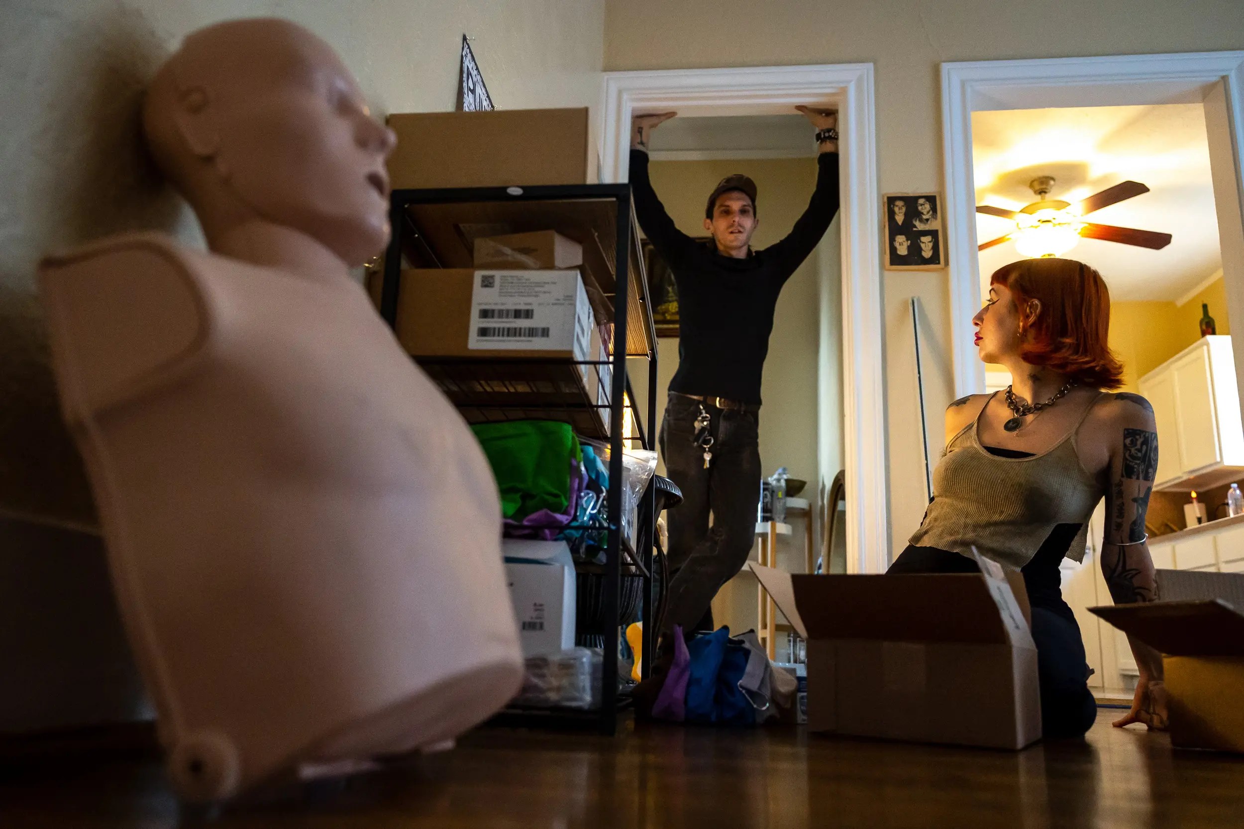 A man wearing black and a ball cap stands in a doorway talking with a woman sitting on the floor. She is wearing a sleeveless top and has tatoos on her left arm. In the foreground is a CPR dummy.