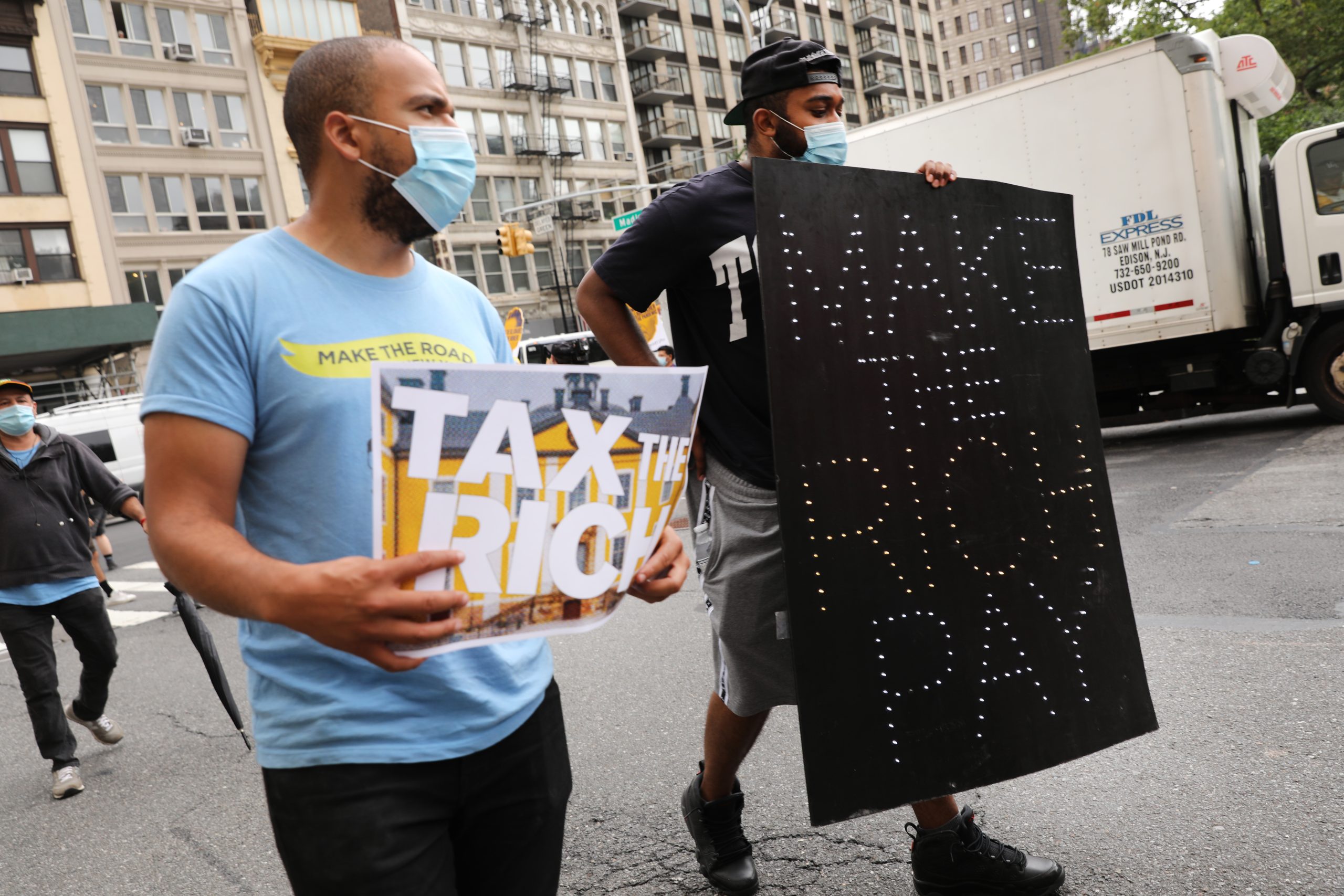 Two men wearing blue surgical masks to protect from covid hold signs that say "Tax the Rich" and "Make the Rich Pay." They're walking outside with people behind them.