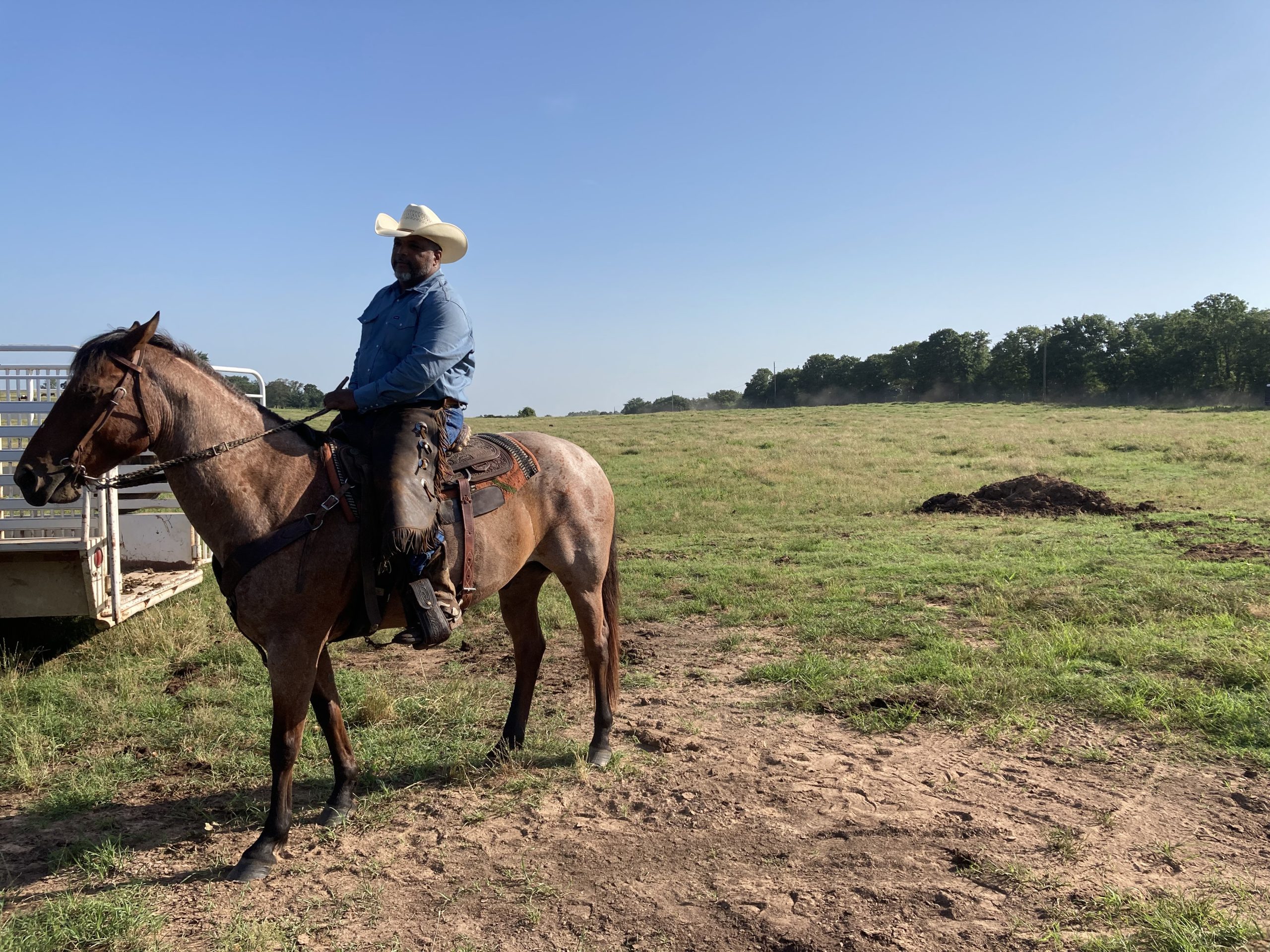 Nate dressed in a cowboy hat, blue hat and pants sits on a horse. His large farm with tall trees is in the background.