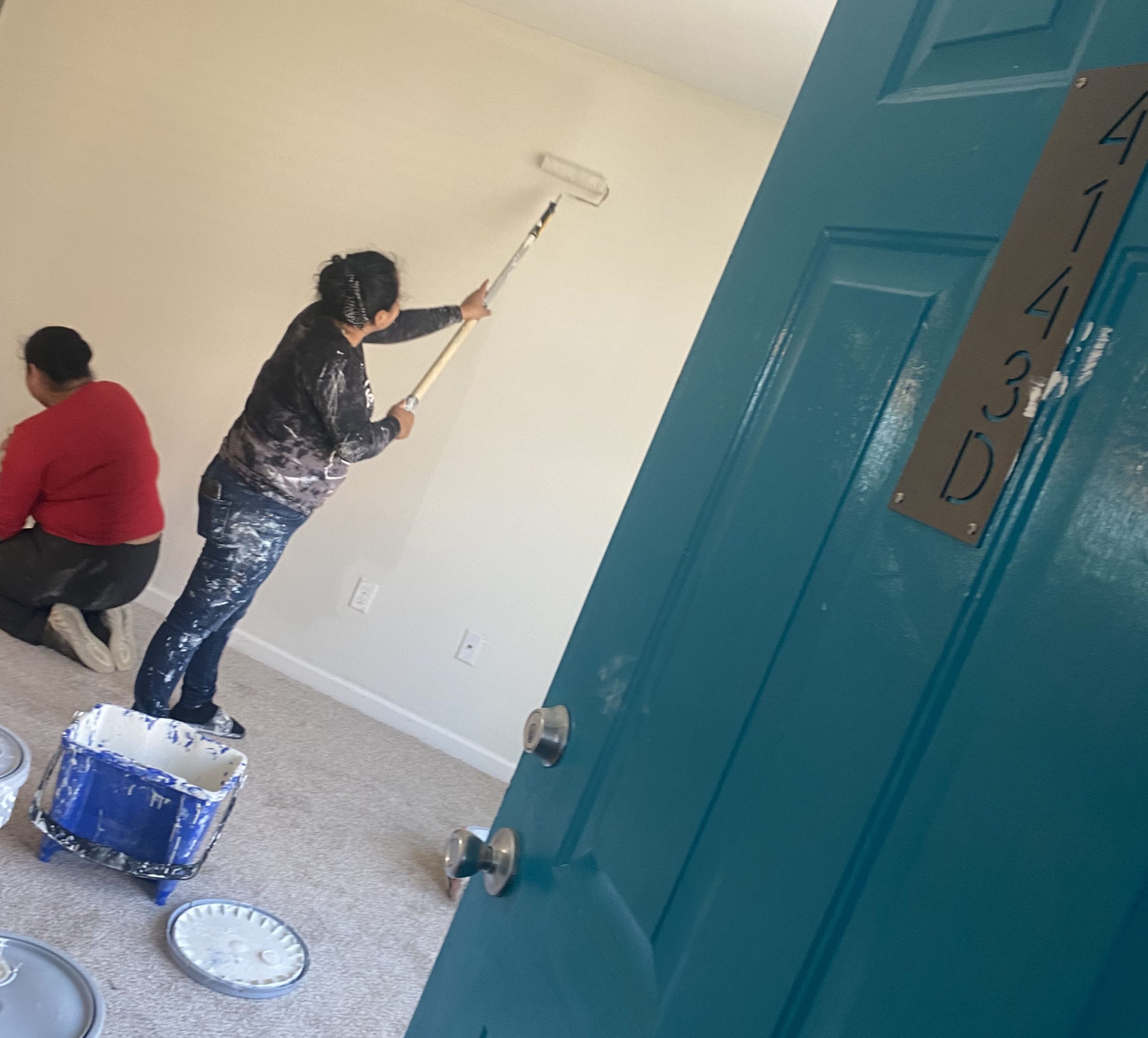 Disaster restoration worker Jenny, dressed in a black shirt and jeans, and her sister Abigail, dressed in an orange shirt and black pants, can be seen through a teal doorway, painting a vacant apartment at the Mayfair Apartment Homes in New Orleans with rollers and brushes, facing away from the camera. The right side of Jenny’s body is covered in splotches of white paint.