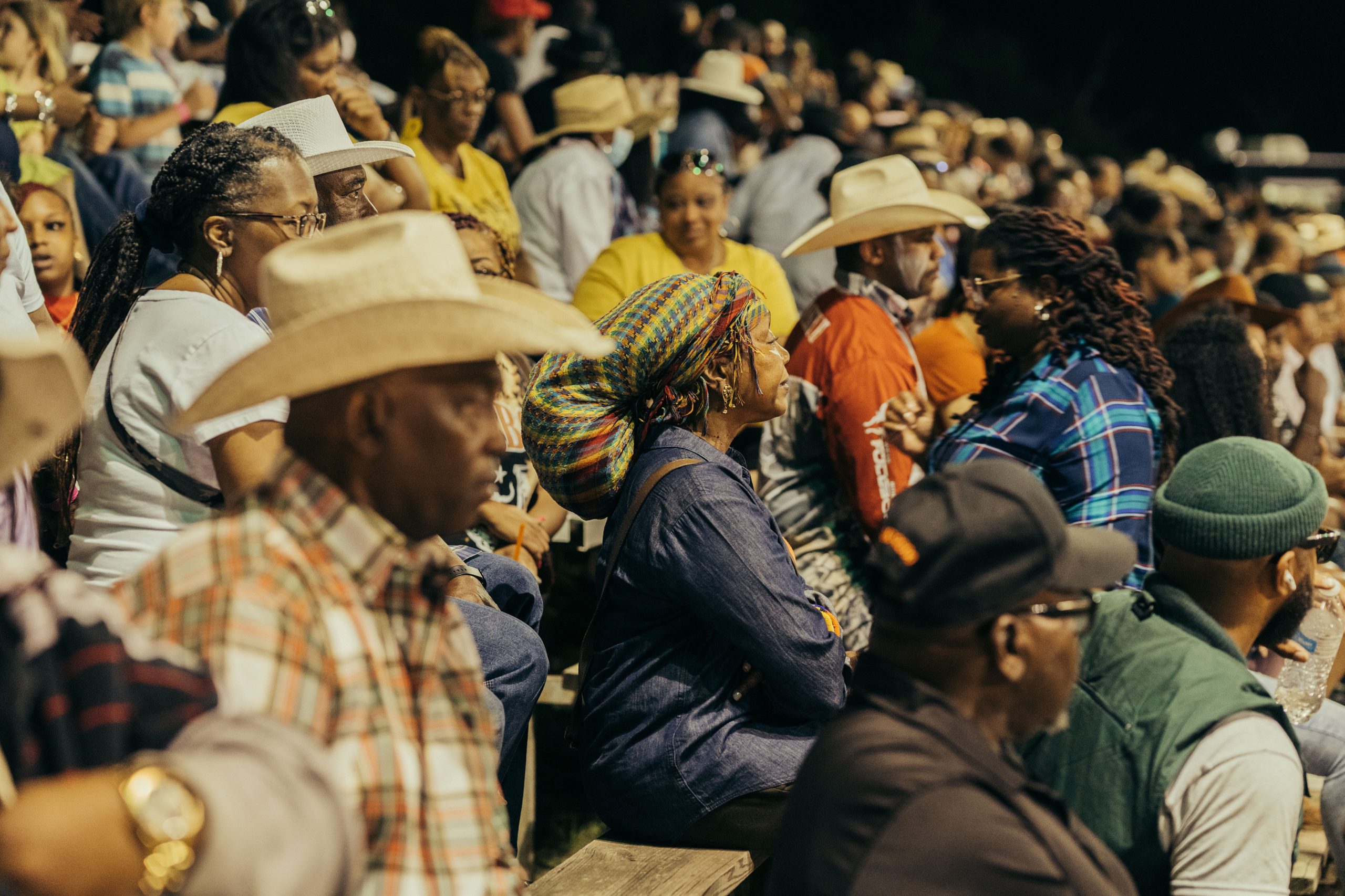 Attendees, some wearing cowboy hats, sit in the stands watching the rodeo.