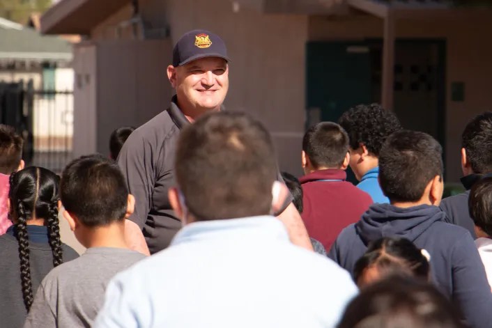 A school resource officer for Charles W. Harris School in Phoenix, greets children at the school during his shift. He wears a black cap and a short-sleeved shirt. He faces children who walk towards him.